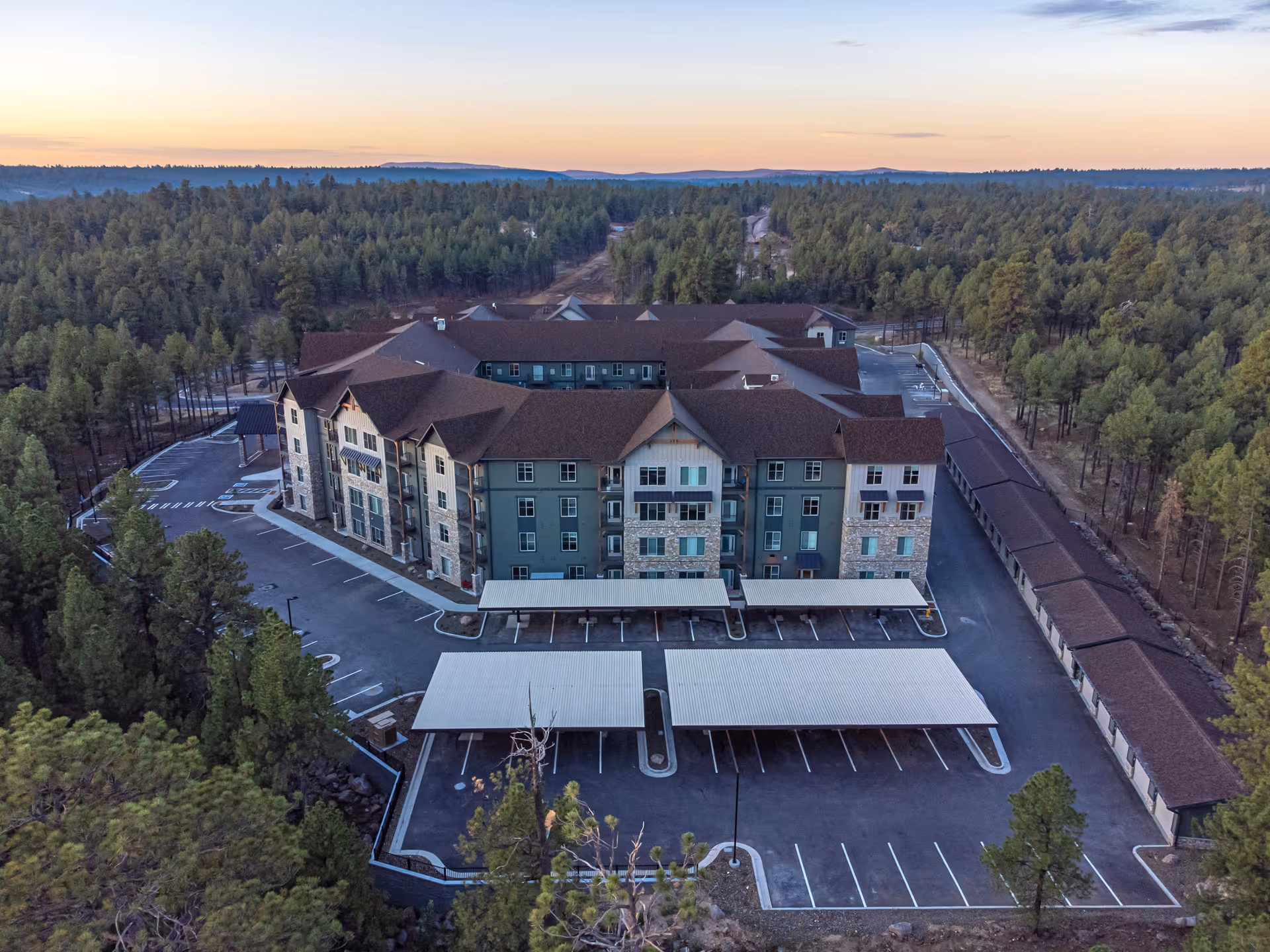 Aerial view of The Bluffs of Flagstaff Senior Living facility surrounded by dense forest. The multi-story building has a brown roof and greenish exterior walls with stone accents. There are covered parking spaces and an adjacent long covered structure, possibly storage or additional parking. The scene is captured during sunset with a clear sky and distant hills in the background.