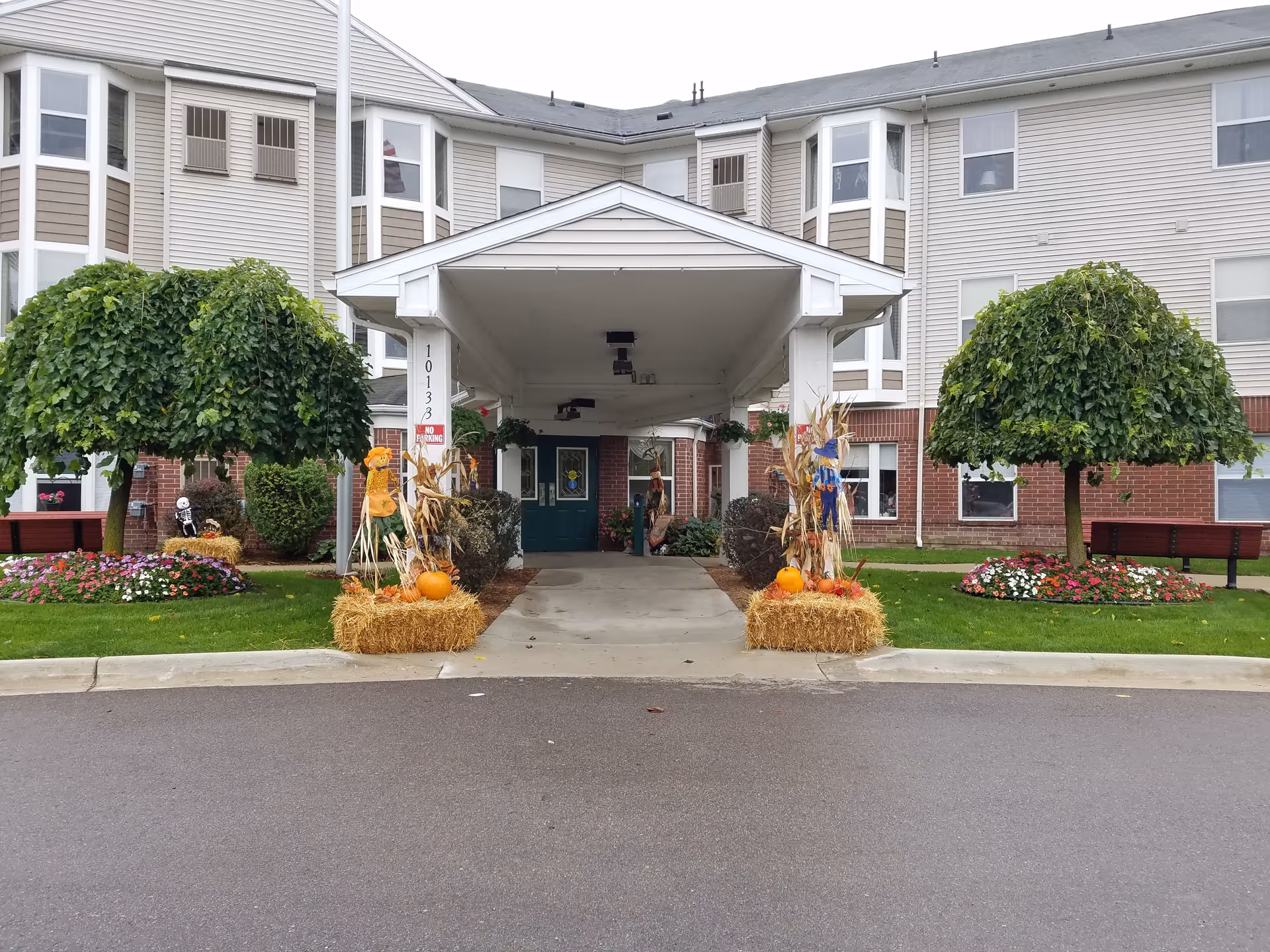 Front entrance of a senior living building with a covered drop-off, fall decorations of hay bales and pumpkins, and landscaped trees and flowerbeds.