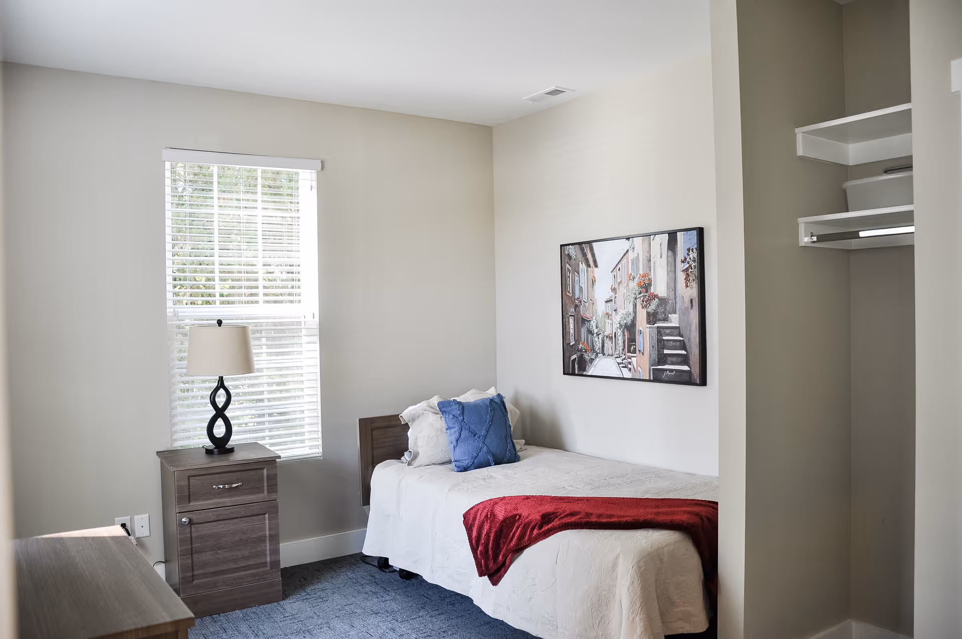 A small bedroom with a single bed covered in a white bedspread and a red throw blanket. There are two pillows on the bed, one white and one blue. A nightstand with a lamp featuring a twisted black base is positioned next to the bed under a window with white blinds. A framed painting of a street scene hangs on the wall above the bed. To the right, there is an open closet with shelves and a hanging rod.