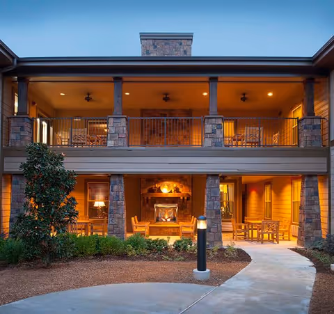 Exterior view of a two-story senior living facility building at dusk with warm lighting. The lower level features a covered patio area with a lit fireplace, wooden chairs, and tables. The upper level has a balcony with railings and ceiling fans. There are shrubs and a tree in front of the building, and a curved concrete walkway leading to the entrance.