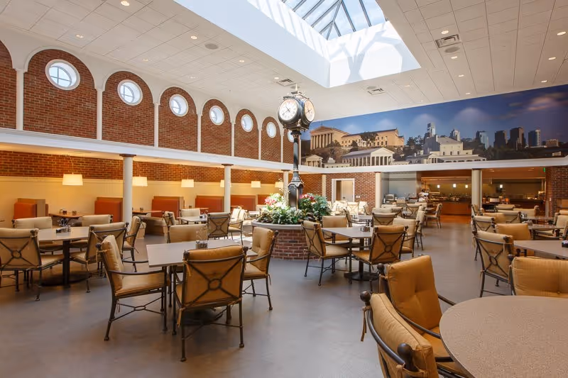 Spacious dining room with multiple tables and cushioned chairs arranged around a central clock and planter beneath a large skylight.