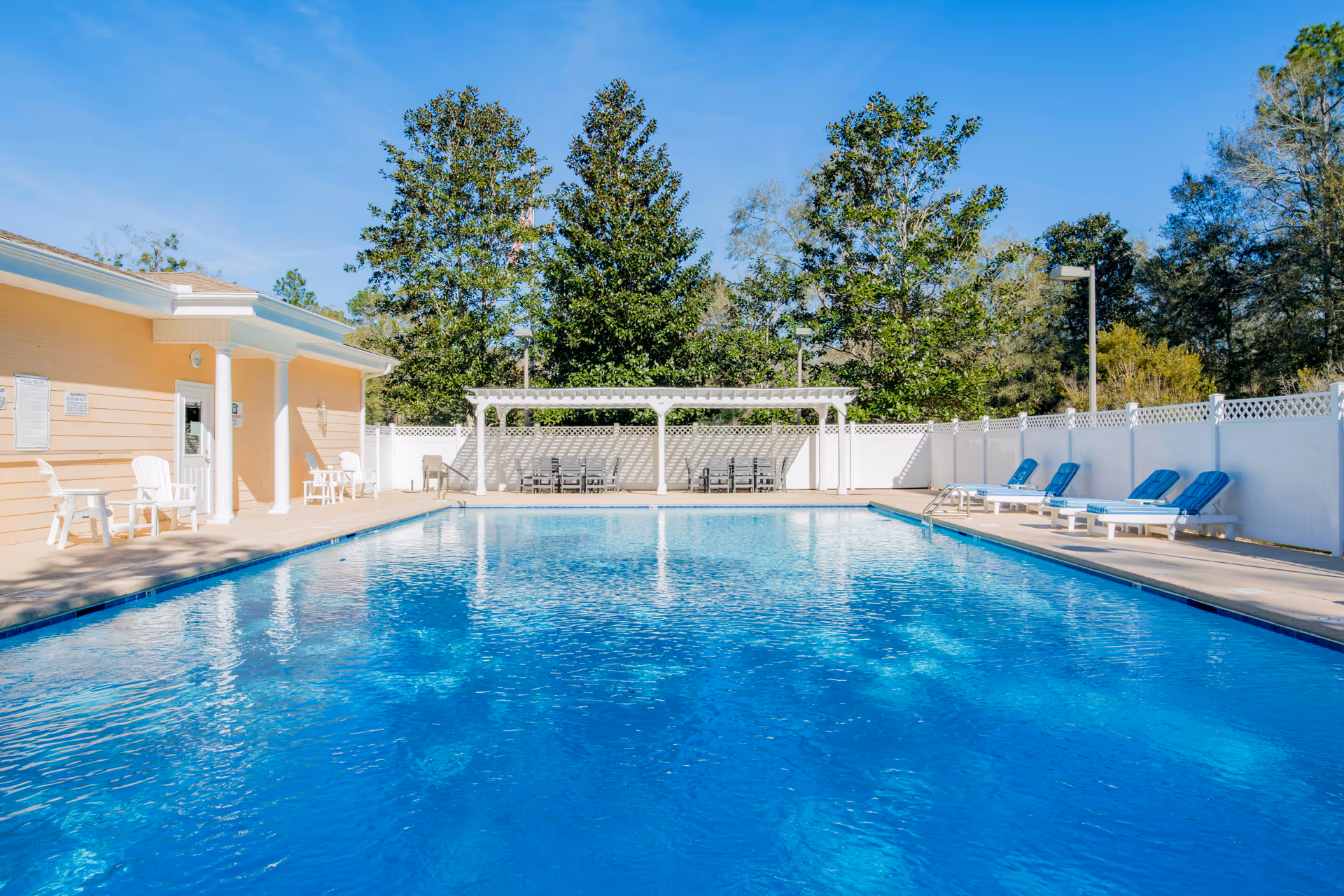 Outdoor swimming pool with clear blue water surrounded by a concrete deck. There are white lounge chairs with blue cushions on the right side and white chairs on the left side near a light yellow building with white columns. In the background, there is a white pergola with chairs underneath and tall green trees behind a white fence.