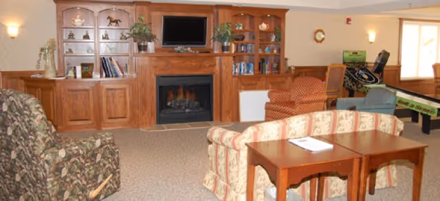 A cozy common living room with a wood-paneled built-in fireplace and TV, surrounded by upholstered sofas, chairs, and small tables.