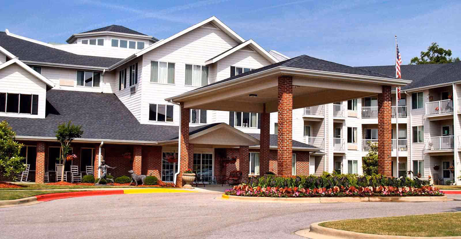 Exterior view of Asher Point Independent Living of Hoover, showing a large building with white siding and brick accents, a covered entrance supported by brick columns, landscaped flower beds, and an American flag on a flagpole.