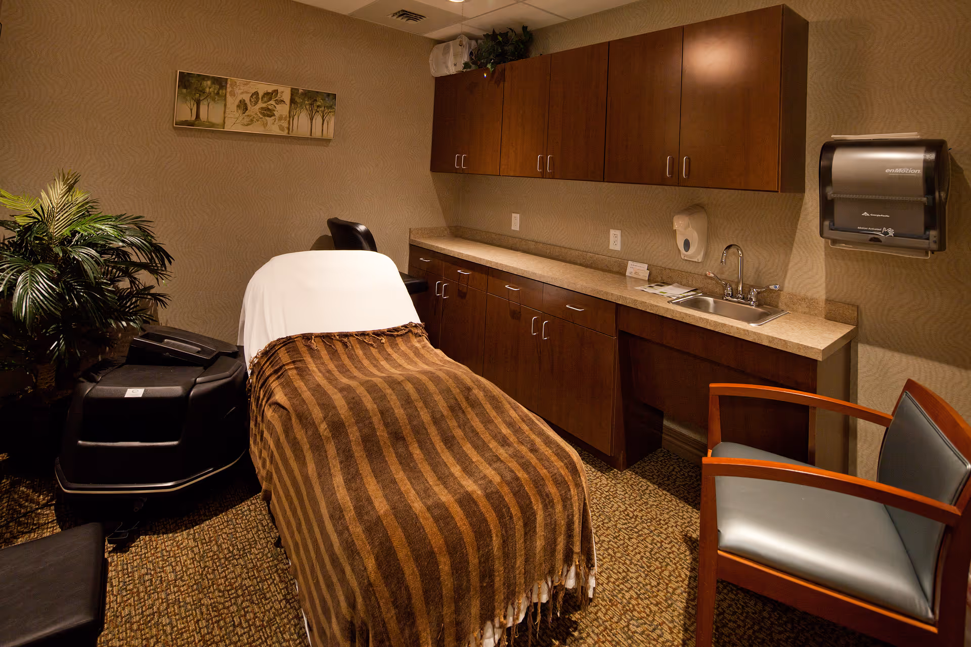 A cozy treatment room with a massage or therapy table covered with a brown striped blanket and a white sheet. The room features wooden cabinets along one wall with a countertop and a small sink. There is a comfortable chair with wooden arms and a potted plant in the corner. The walls have a subtle textured pattern and a framed artwork with tree and leaf designs hangs above the table.