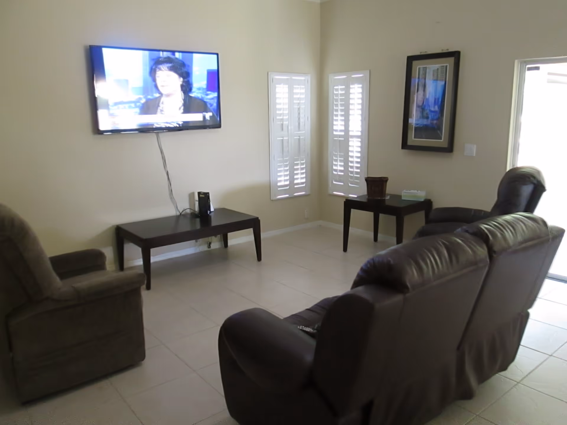 A living room with a wall-mounted flat-screen TV showing a woman speaking. The room has a brown leather sofa, a brown recliner chair, a dark wooden coffee table under the TV, and a side table with a tissue box and decorative item. There are two windows with white shutters and a framed picture on the wall.