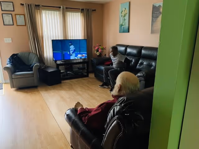 A living room with two elderly men seated on black leather sofas watching a baseball game on a flat-screen TV. The room has light wooden flooring, beige walls with framed pictures, a gray armchair, and a window with vertical blinds and curtains.