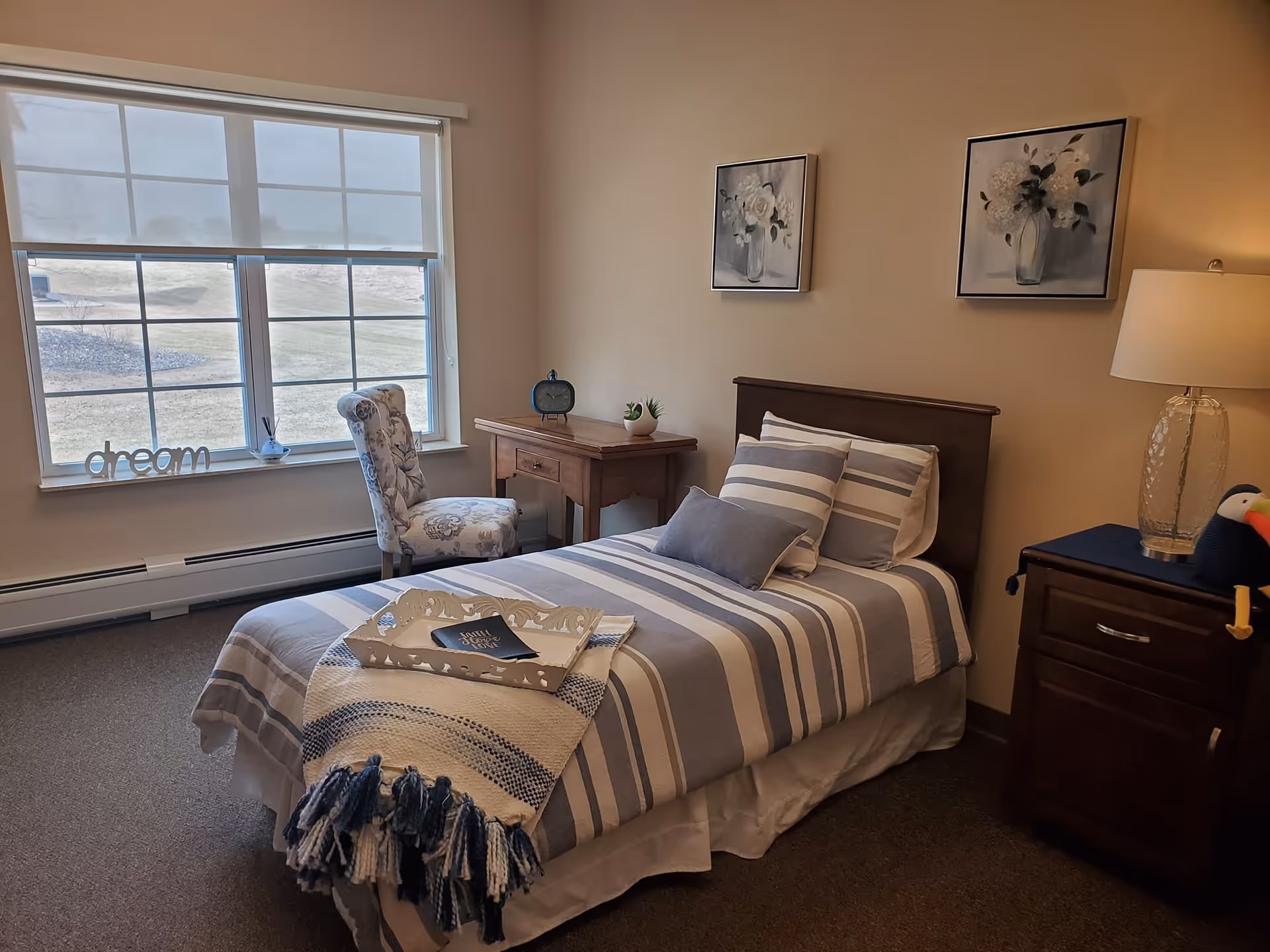 Neat single bed in a decorated bedroom with striped linens, a nightstand, desk, chair, and a large window.