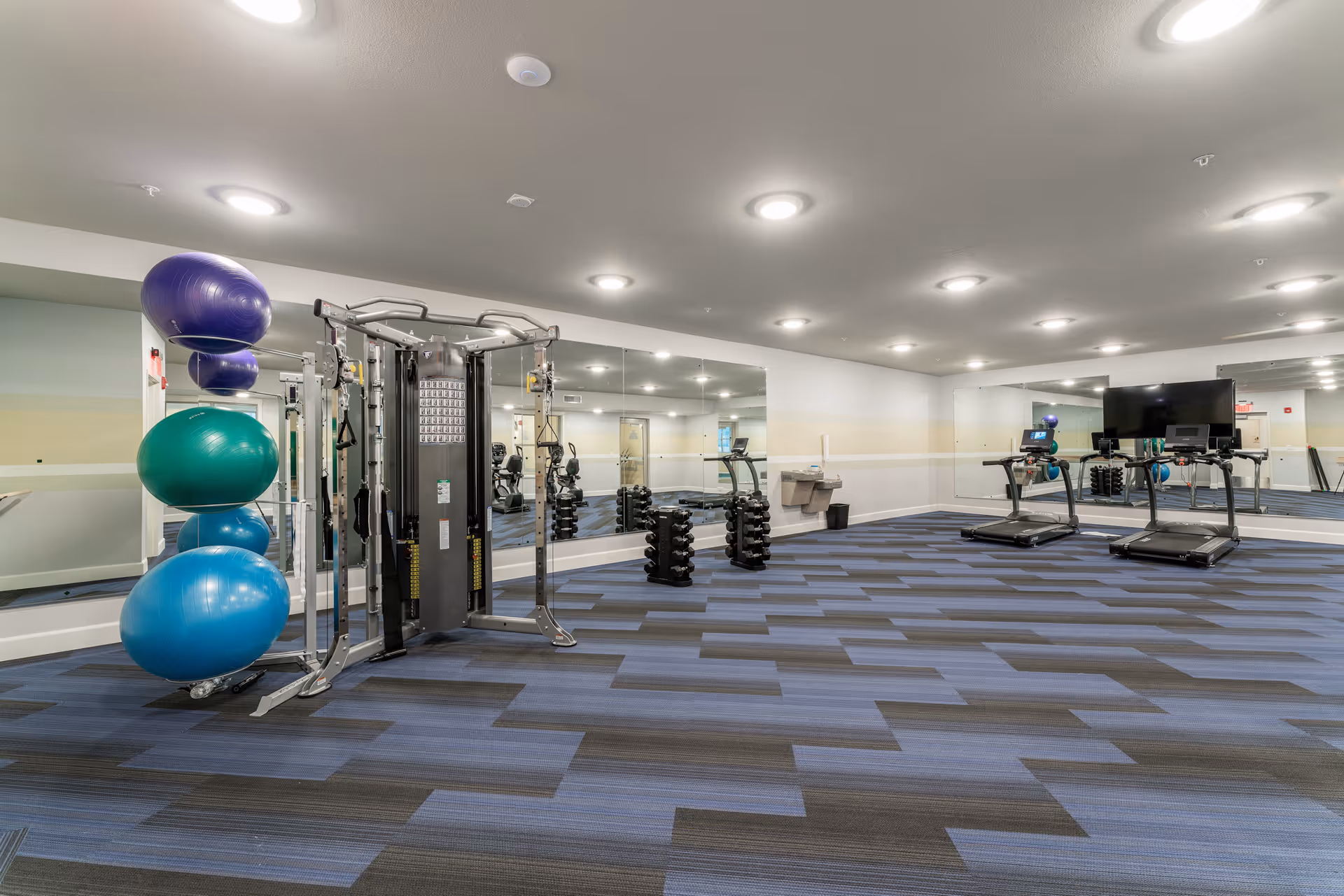 A senior living facility fitness room with exercise equipment including a multi-functional weight machine, two treadmills, several dumbbell racks, and large exercise balls stacked on a rack. The room has blue and gray patterned carpet, white walls with a beige stripe, and large mirrors covering two walls. Ceiling lights illuminate the space.