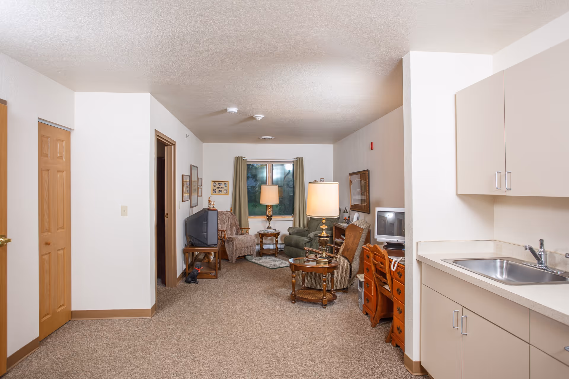 Interior view of a senior living facility room showing a small kitchenette area with a sink and cabinets on the right, and a living area with armchairs, a side table with a lamp, a TV on a stand, and a window with curtains in the background. The room has beige carpet and white walls with wooden trim and doors.