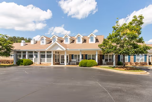 Front exterior view of a single-story senior living facility building with a beige roof, multiple dormer windows, white pillars, and a paved driveway. There are green bushes and trees around the building under a partly cloudy blue sky.