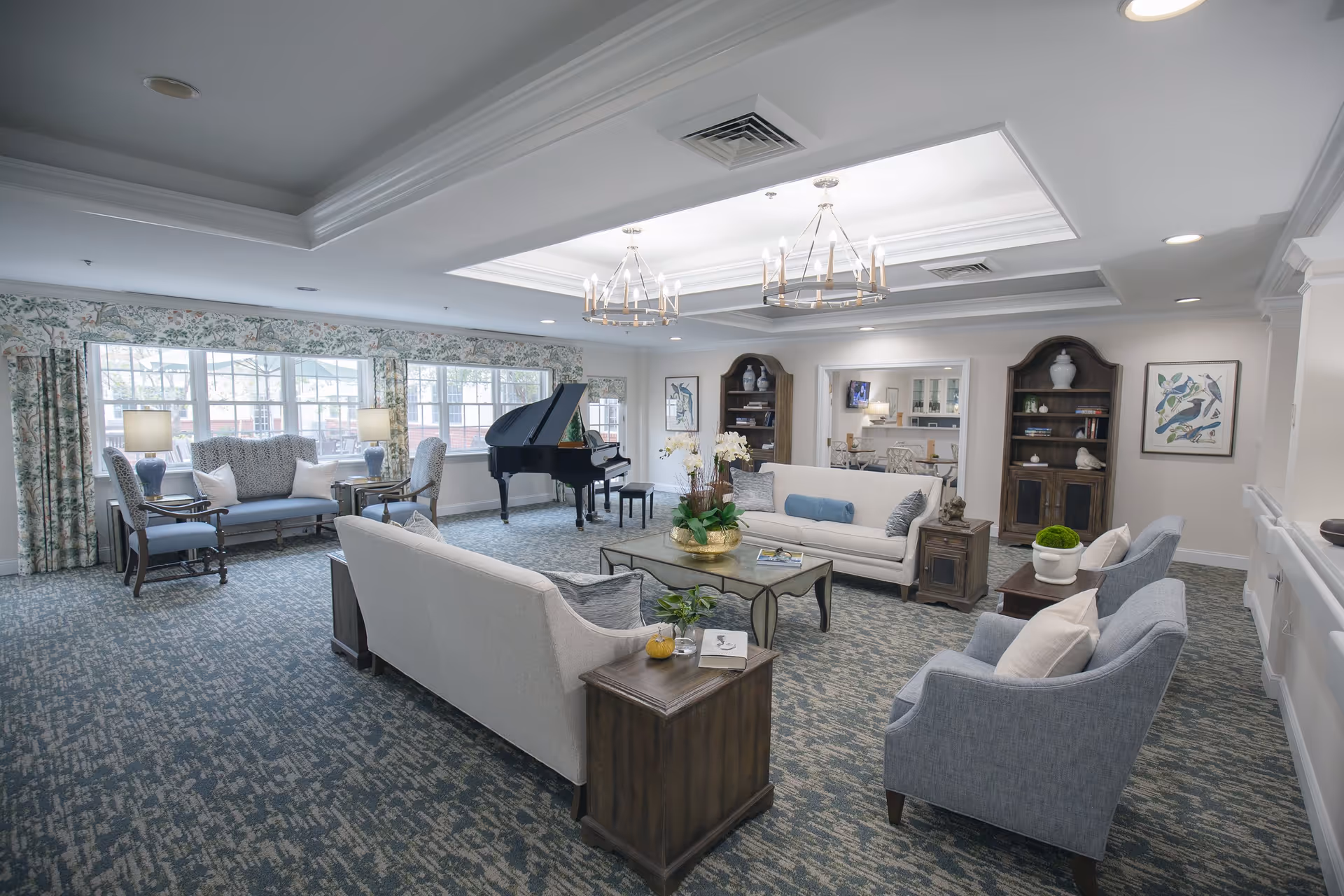 A spacious and well-lit living room area with multiple seating options including sofas and armchairs arranged around a central coffee table. The room features large windows with floral curtains, two chandeliers, a grand piano in the corner, bookshelves with decorative items, and framed bird artwork on the walls.