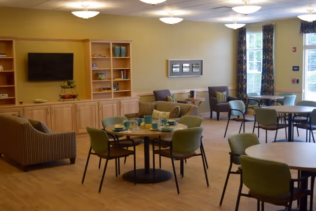 A spacious and well-lit common area with multiple round tables and green cushioned chairs arranged for dining or socializing. There are sofas and armchairs near a wooden entertainment center with a flat-screen TV mounted on the wall. Large windows with patterned curtains allow natural light to fill the room.