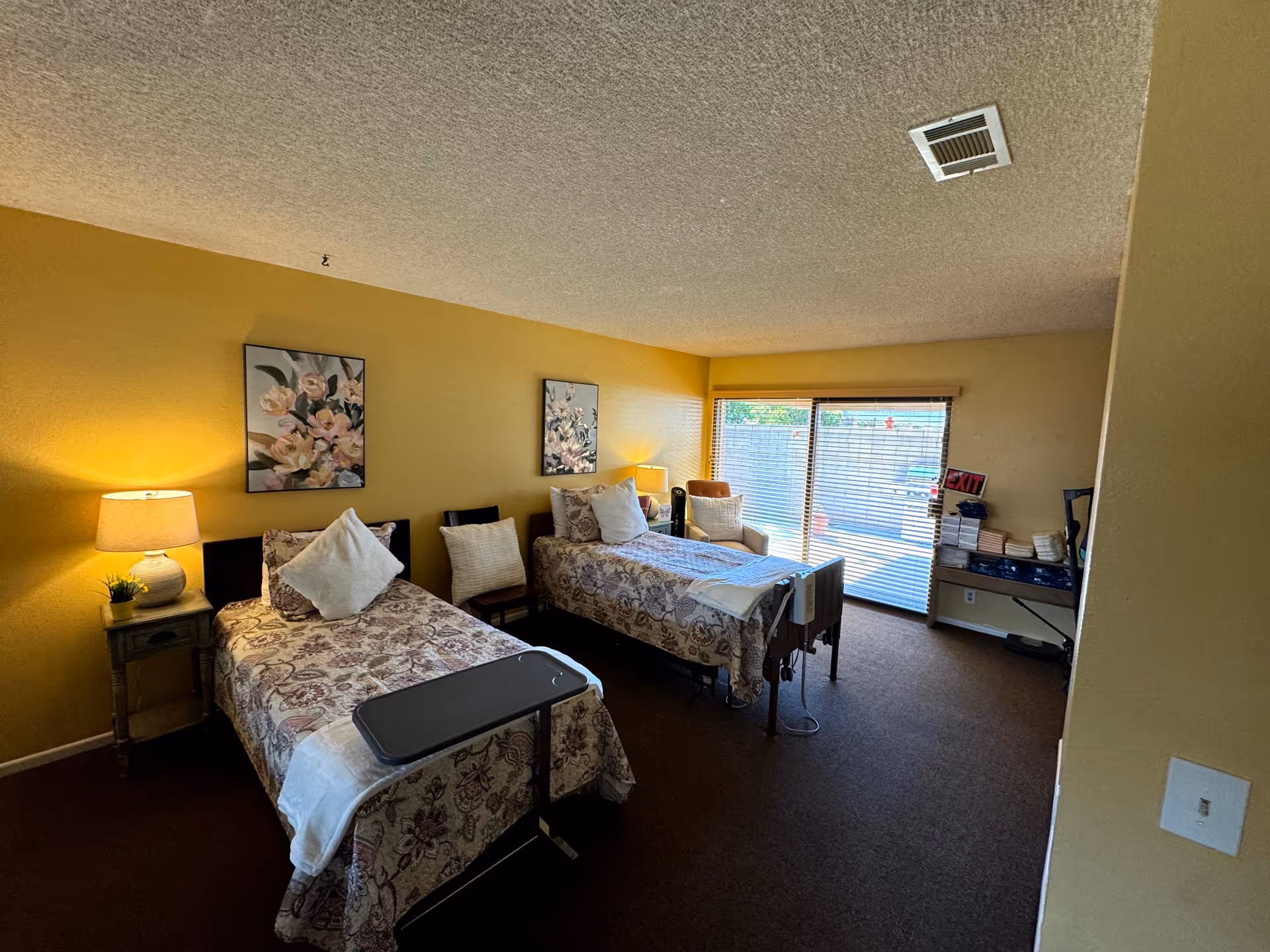 A senior care facility bedroom with two single beds, each with floral bedspreads and pillows. There is a small bedside table with a lamp and a small plant between the beds. Two floral paintings hang on the yellow wall above the beds. A chair is placed between the beds. The room has a large window with blinds, letting in natural light. A small desk with books and an exit sign is visible near the window.
