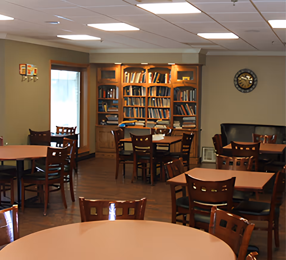A communal room with multiple wooden tables and chairs arranged for seating. In the background, there is a large wooden bookshelf filled with books. The room has beige walls, a clock on the wall, and a window letting in natural light.