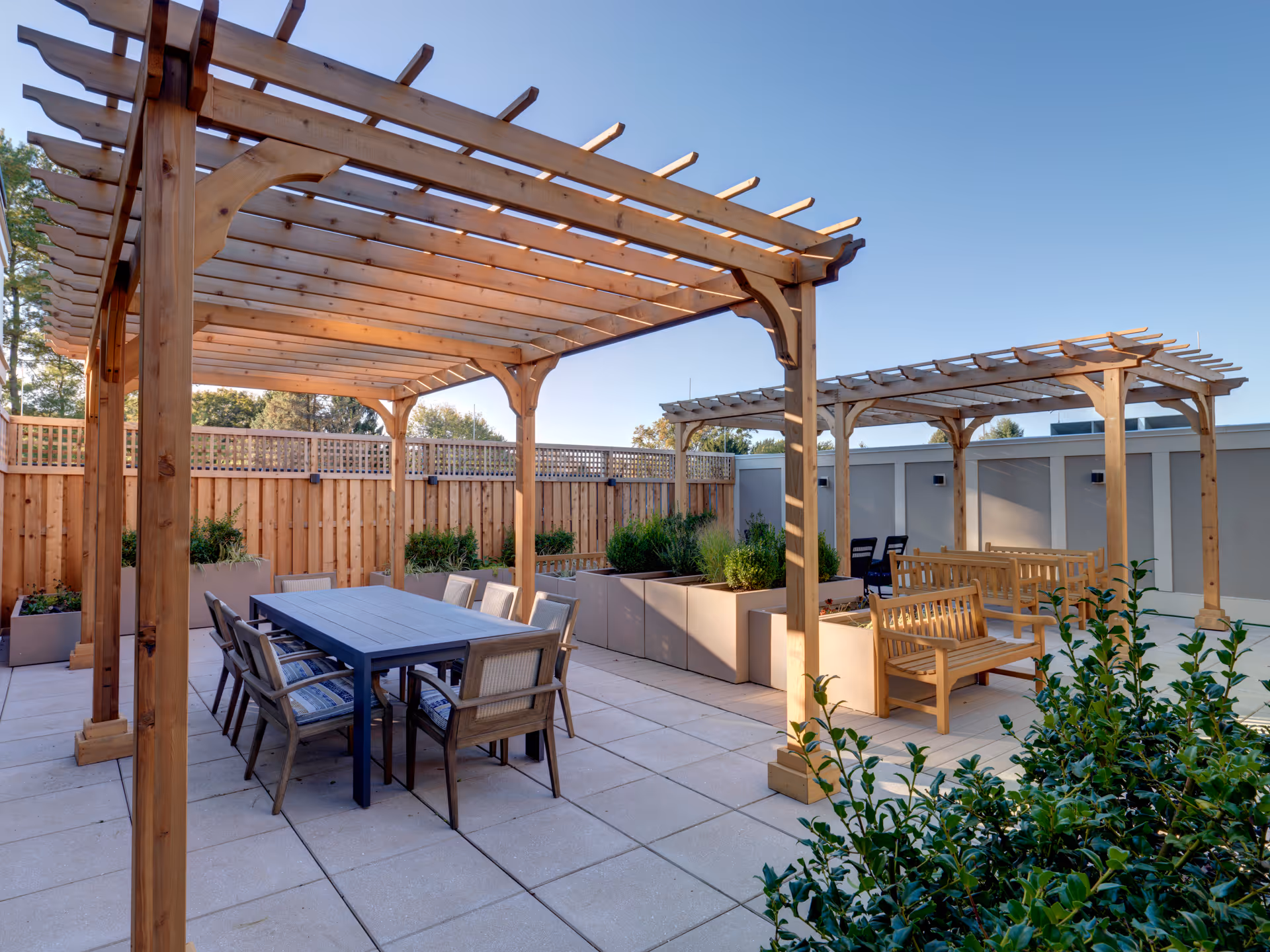 Outdoor patio area with two wooden pergolas, one covering a dining table with chairs and the other covering wooden benches. The space is surrounded by a wooden fence and planter boxes with greenery under a clear blue sky.