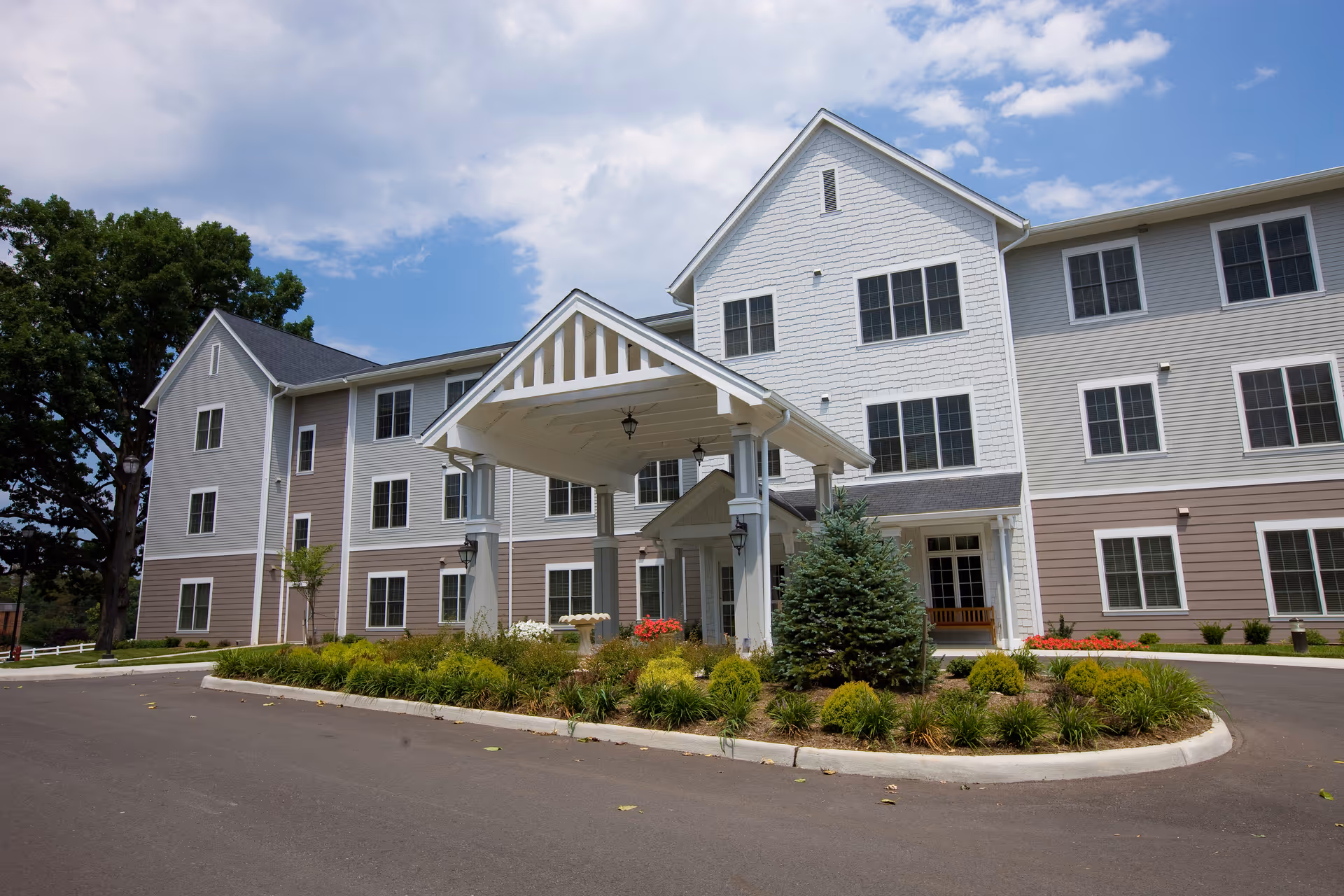 Exterior view of a multi-story senior living facility building with a covered entrance, landscaped greenery, and a paved driveway under a partly cloudy sky.