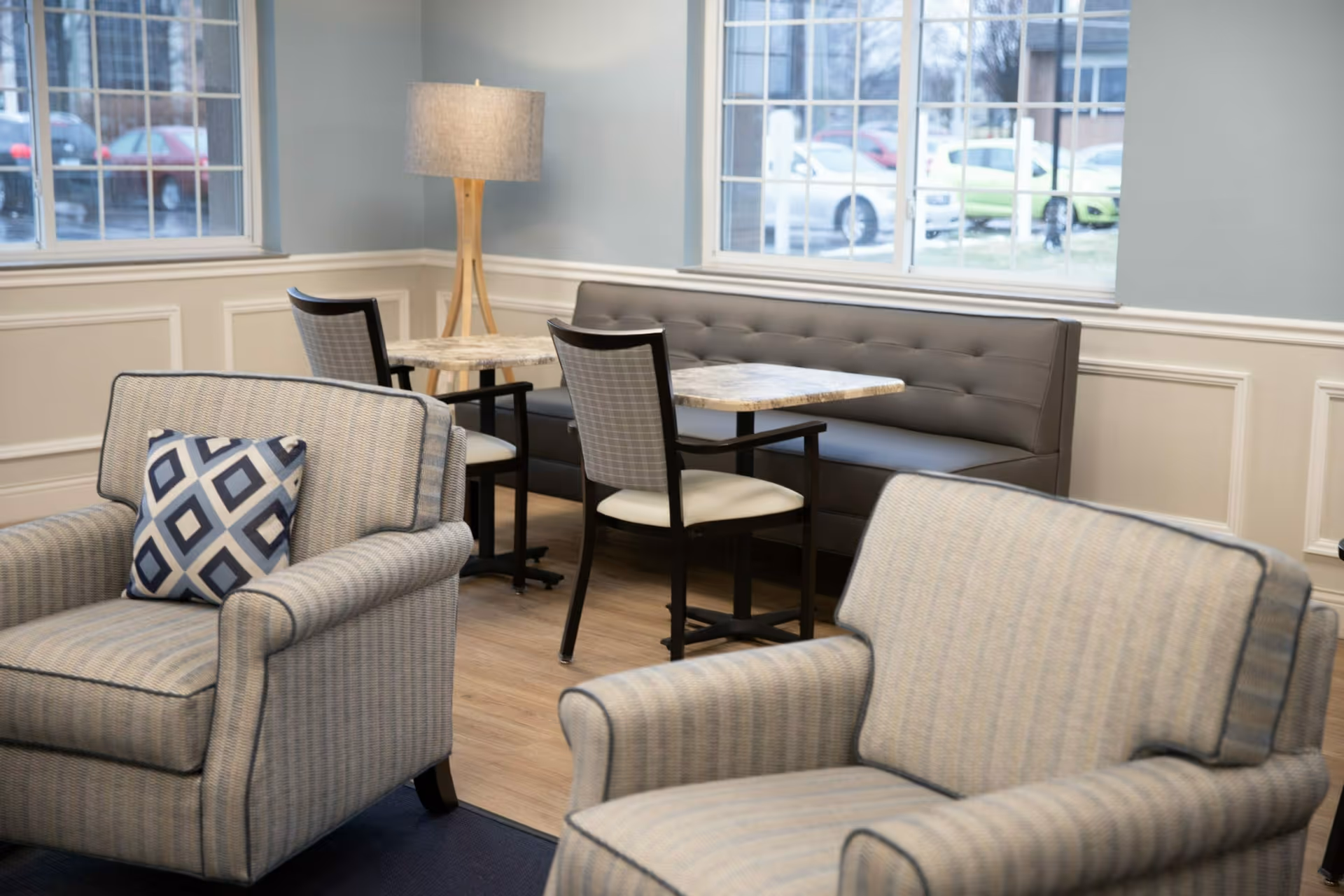 A cozy seating area in a senior living facility with two upholstered armchairs featuring a patterned cushion, a small dining area with two tables, chairs, and a cushioned bench along the wall, illuminated by a floor lamp near large windows showing parked cars outside.