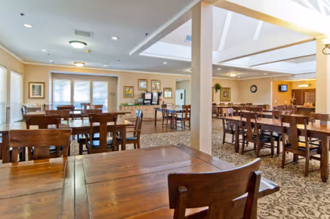 A spacious dining room with multiple wooden tables and chairs arranged neatly. The room has large windows allowing natural light to enter, beige walls adorned with framed pictures, and a carpeted floor with a patterned design. There is a beverage station against the far wall and ceiling lights providing additional illumination.