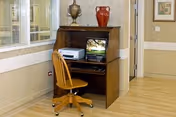 A small wooden computer workstation with a monitor, printer, and rolling wooden chair against a wainscoted hallway wall, topped with decorative vases.