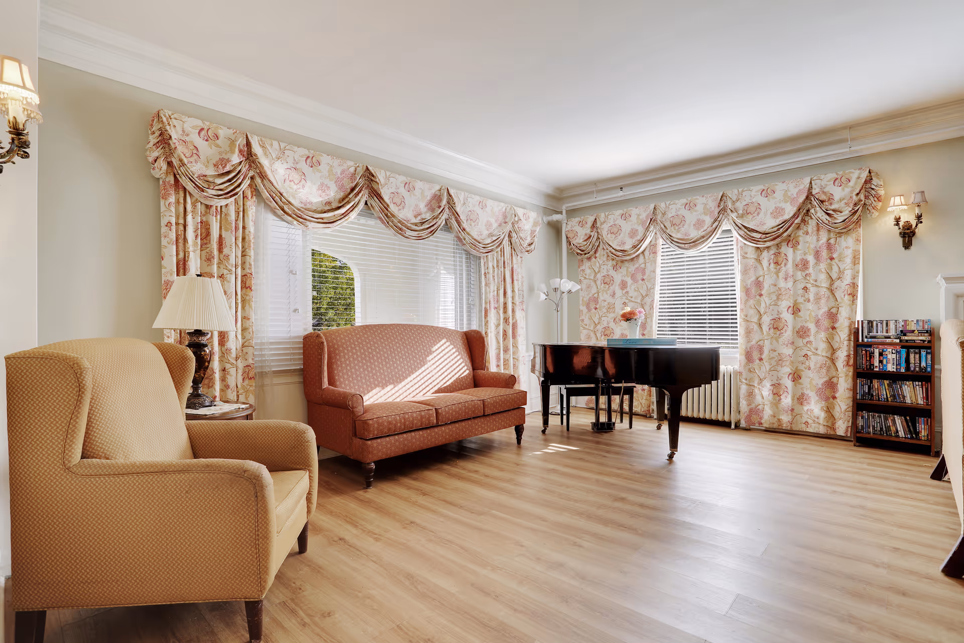 A bright living room with wooden flooring featuring a beige armchair, a red patterned loveseat, a black grand piano, floral curtains on two windows, a small bookshelf filled with DVDs, and wall-mounted lamps.