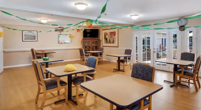 A dining room in Greenwood Assisted Living decorated with green and yellow streamers hanging from the ceiling. The room contains several square tables with chairs around them, a wooden cabinet with a TV on top, a wall clock, and framed artwork on the walls. The floor is wooden, and there are glass doors leading to another room.