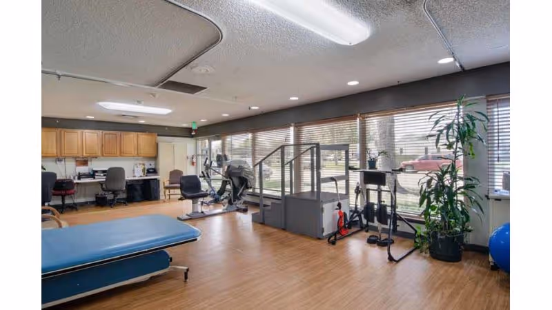 A rehabilitation room with exercise equipment including stationary bikes, a treadmill, and a blue therapy table. The room has large windows with blinds letting in natural light, wooden flooring, and office desks with chairs along the back wall. There are also some plants near the windows.