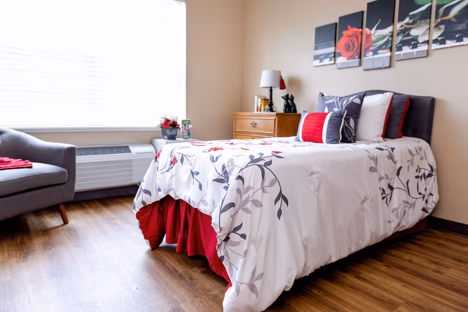 A bright and cozy bedroom with a bed dressed in white and red floral bedding, a gray armchair with a red blanket, a wooden nightstand with a lamp and decorative items, and a large window with blinds letting in natural light. Above the bed, there is a multi-panel wall art featuring a red rose.