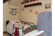 A nursing home bedroom with a single bed, a mobility walker, an upholstered chair, and wall decorations.