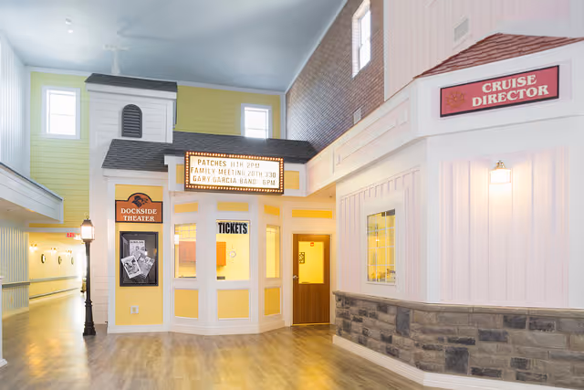 Interior view of a senior living facility hallway with a small theater ticket booth labeled 'Dockside Theater' and a sign above it listing events. To the right is a section labeled 'Cruise Director' with a window and light fixture. The area has light yellow and pink walls, wood flooring, and a high ceiling with windows.