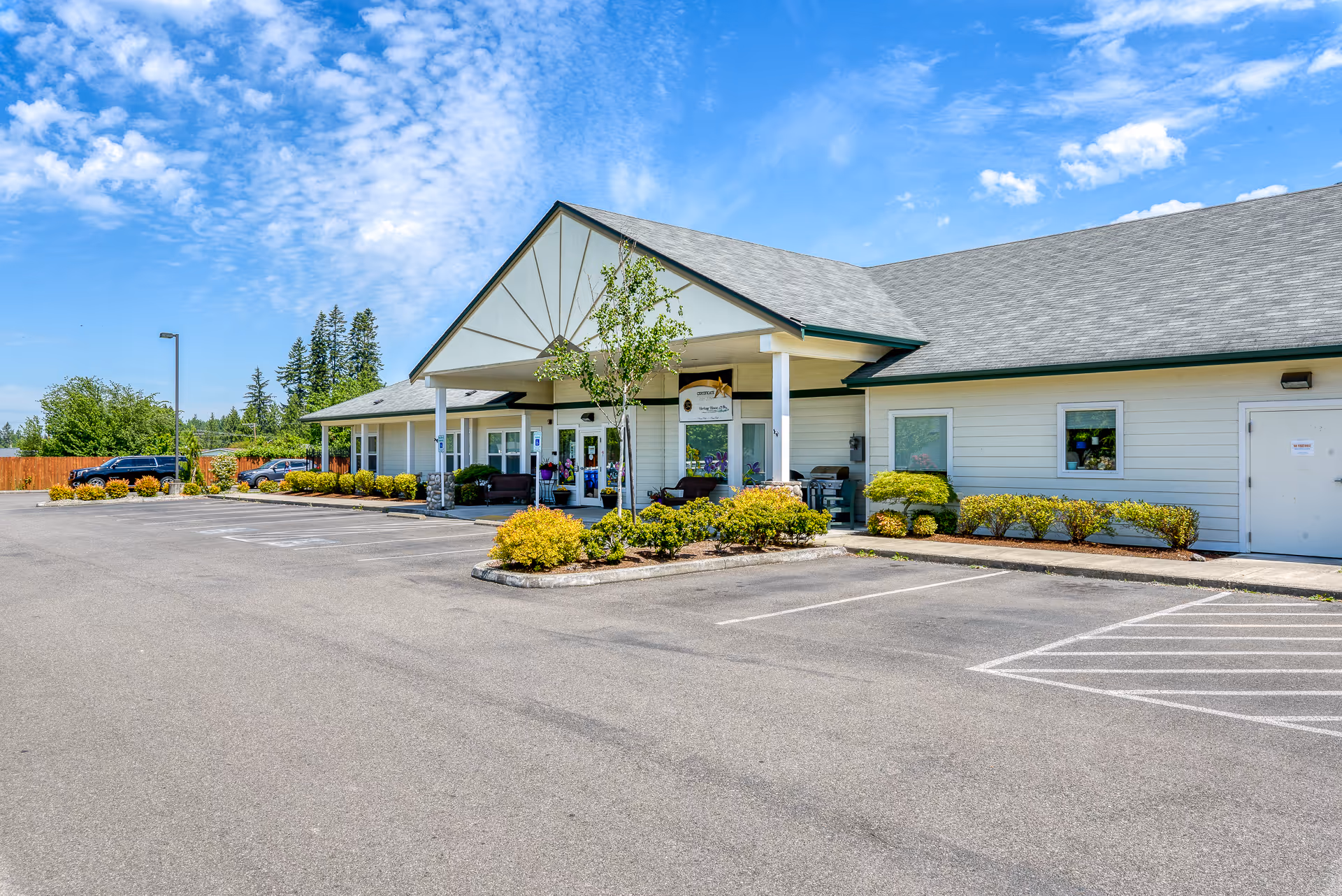 Exterior view of Heritage House Buckley Assisted Living & Memory Care building on a sunny day with a clear blue sky. The building has a light-colored facade with a peaked roof and a covered entrance supported by white pillars. There are shrubs and small trees planted around the entrance and a parking lot with marked spaces in front.