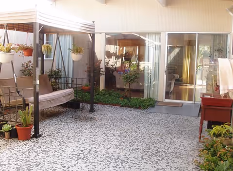 A covered outdoor patio area with a cushioned swing bench, several potted plants, and a small garden bed along the wall. The patio has a speckled floor and is adjacent to a building with large sliding glass doors leading inside.