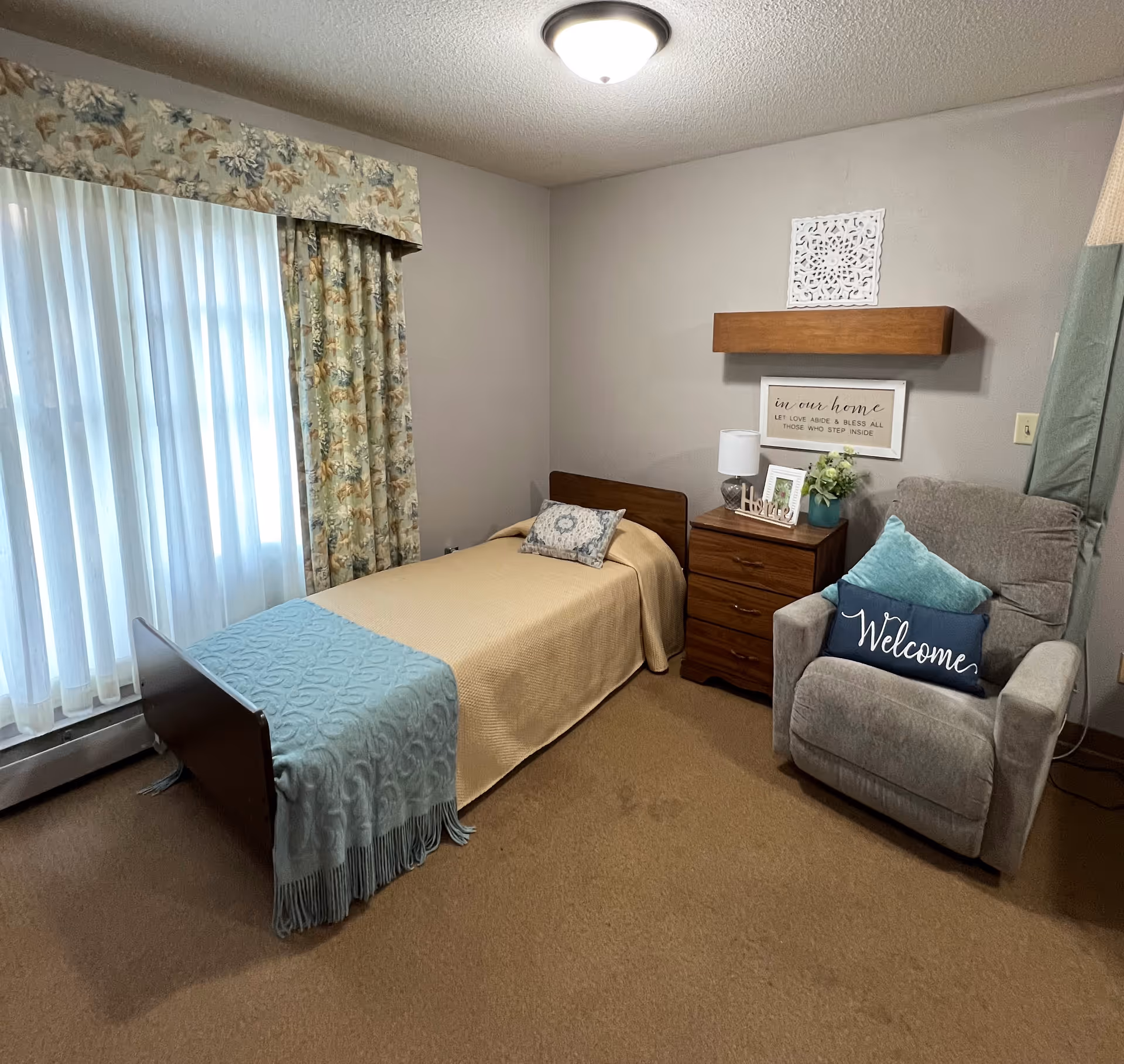 A cozy bedroom in a senior living facility featuring a single bed with a beige blanket and a light blue throw, a wooden nightstand with a lamp, framed photos, and a small plant. Next to the nightstand is a comfortable gray recliner chair with two pillows, one of which says 'Welcome'. The room has beige carpet, floral curtains, and a ceiling light fixture. A decorative wall shelf and framed sign are mounted on the wall above the nightstand.