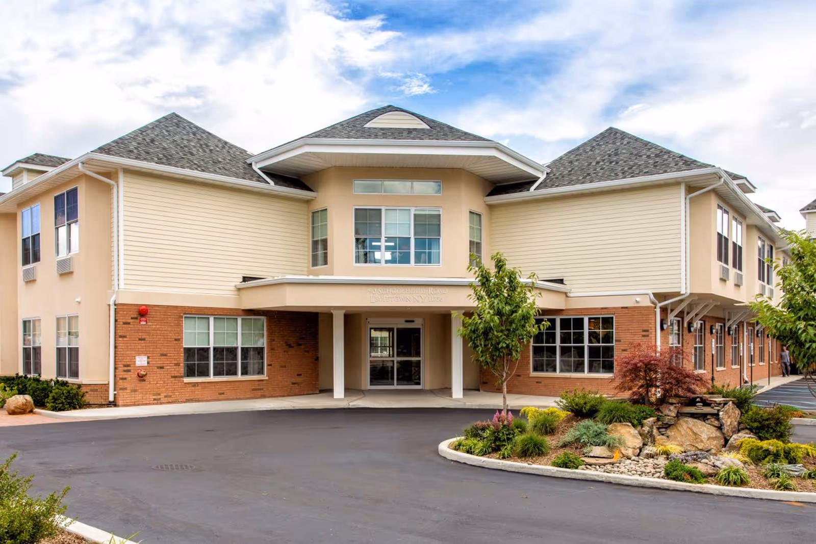 Front exterior view of a two-story senior living facility building with beige and brick walls, multiple windows, a covered entrance, and landscaped greenery with rocks and small trees in front.