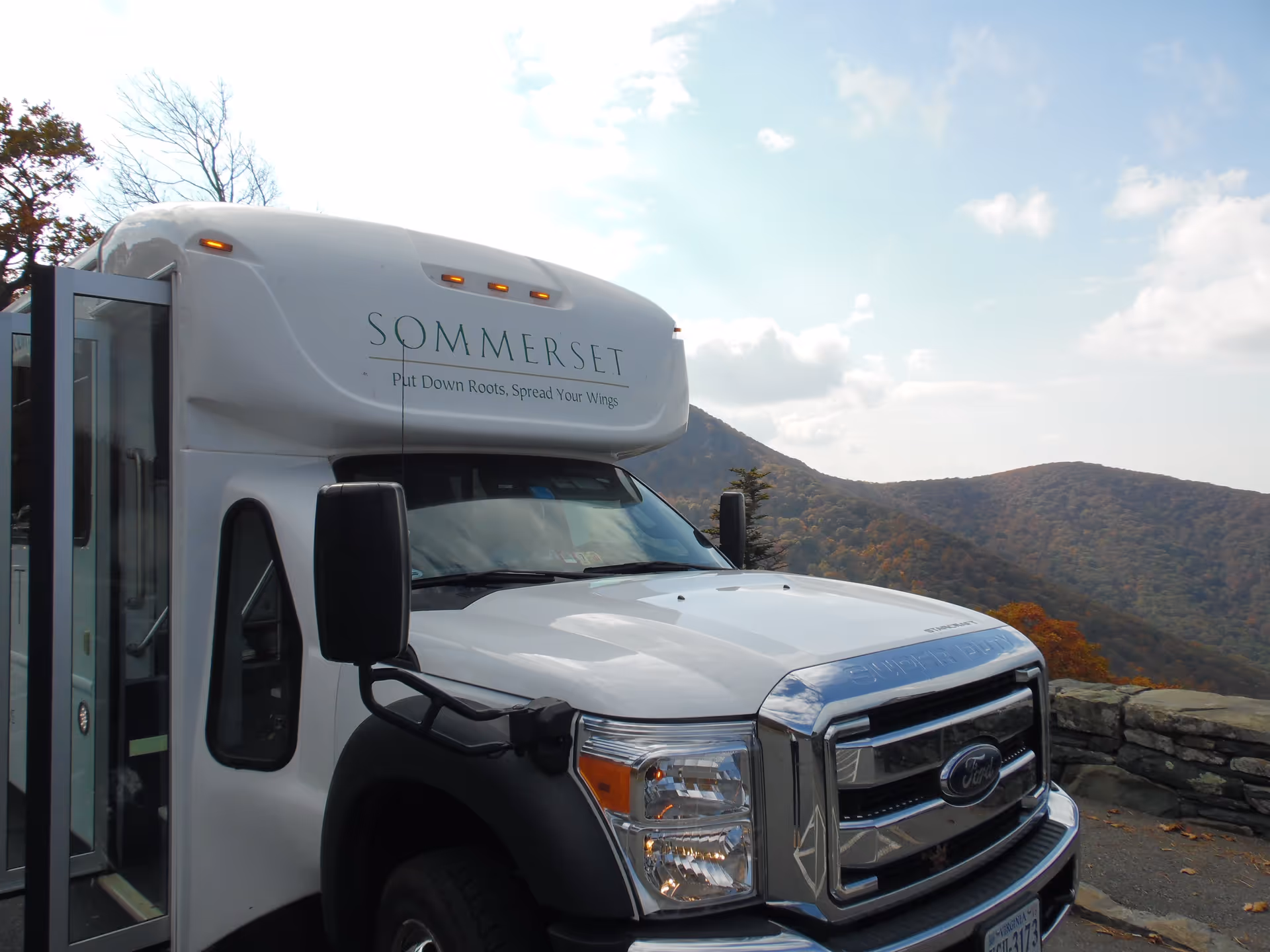 White Sommerset retirement community shuttle parked at a mountain overlook with rolling hills in the background.