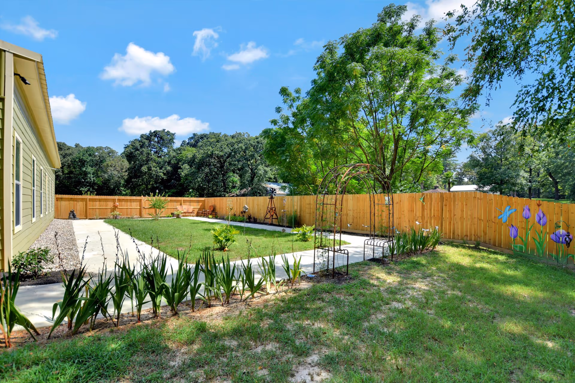 Grassy fenced backyard with a concrete walkway, metal arbor, planted borders, and a yellow building on the left under a blue sky.