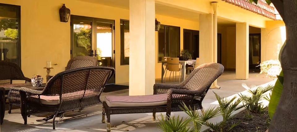 Covered outdoor patio with wicker lounge chairs and small tables along a building walkway.
