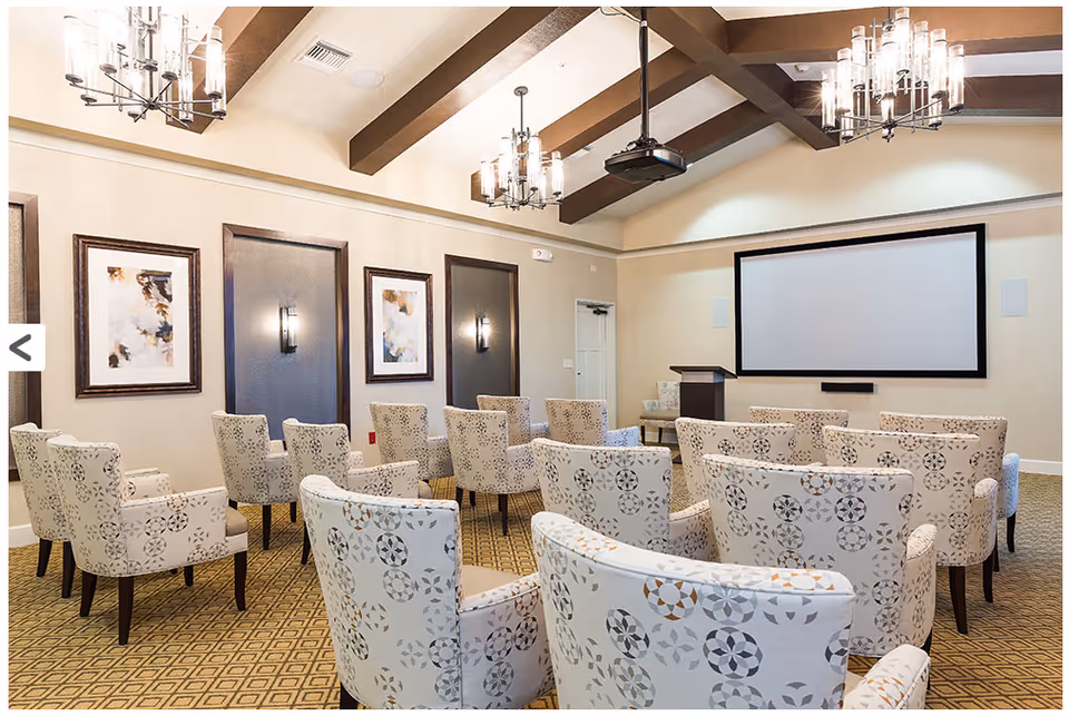 A well-lit meeting or presentation room with patterned armchairs arranged in rows facing a large projection screen and a podium. The room features a high ceiling with exposed wooden beams and modern chandeliers, framed artwork on the walls, and a patterned carpet.
