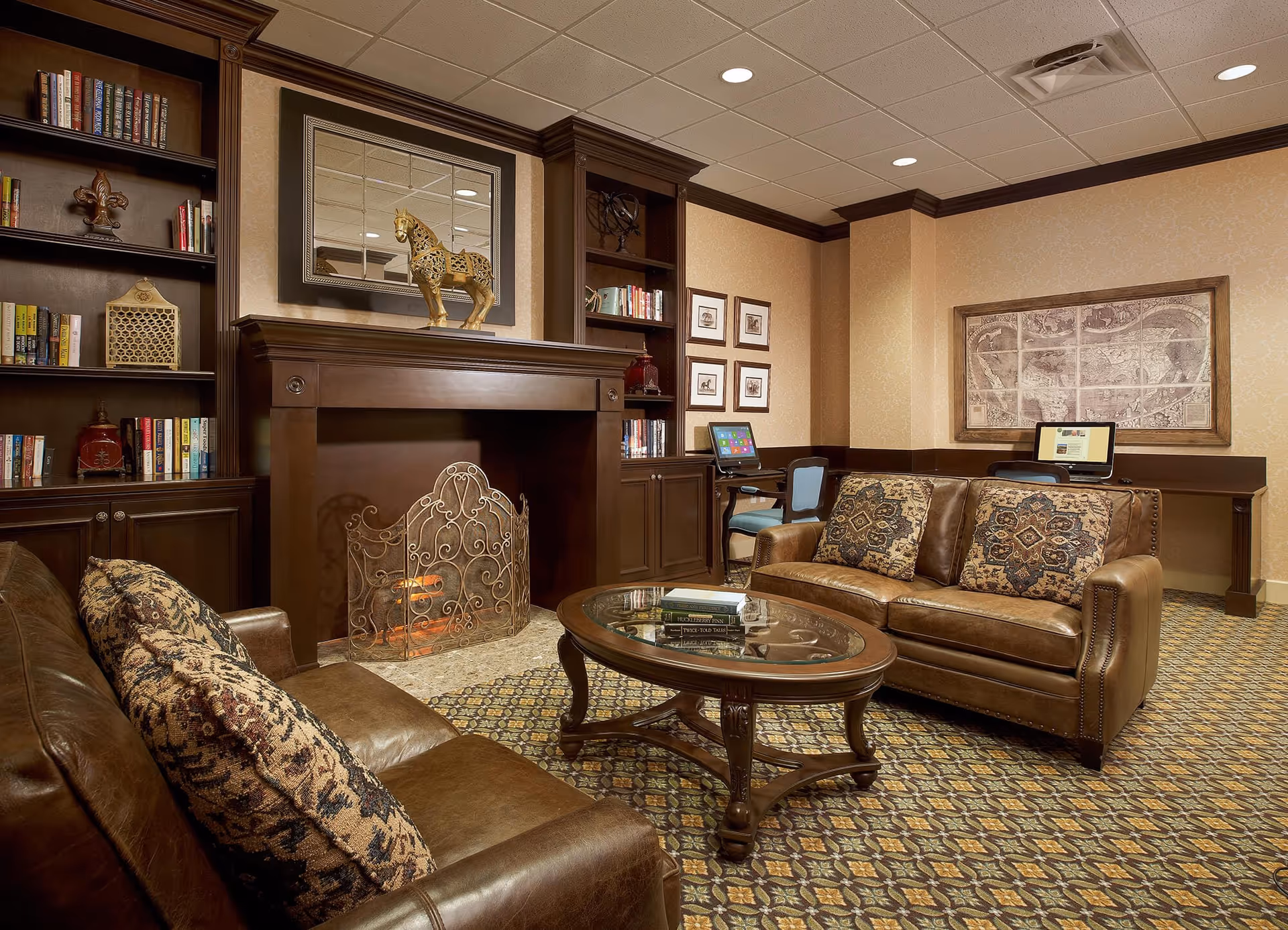 A cozy living room area with two brown leather sofas adorned with patterned cushions, a wooden coffee table with glass top holding books, a decorative fireplace with an ornate screen, built-in dark wood bookshelves filled with books and decorative items, and two computer stations against the wall with framed artwork above.