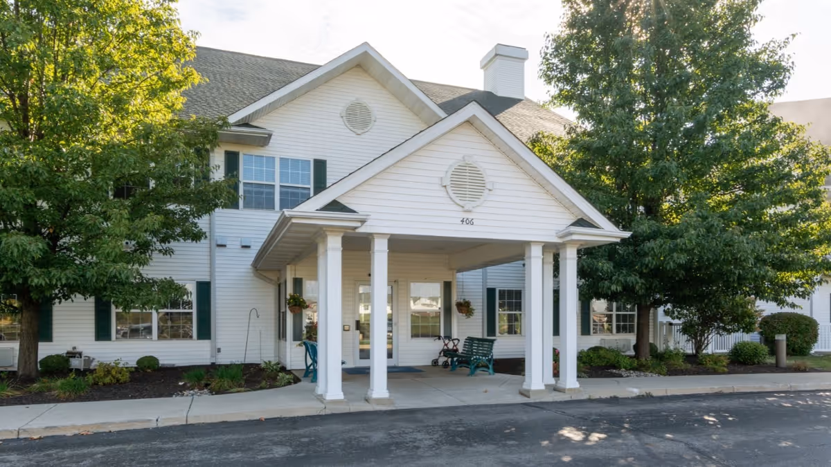 Exterior view of a white two-story senior living facility building with green shutters, a covered entrance supported by four white columns, benches, and trees on either side.