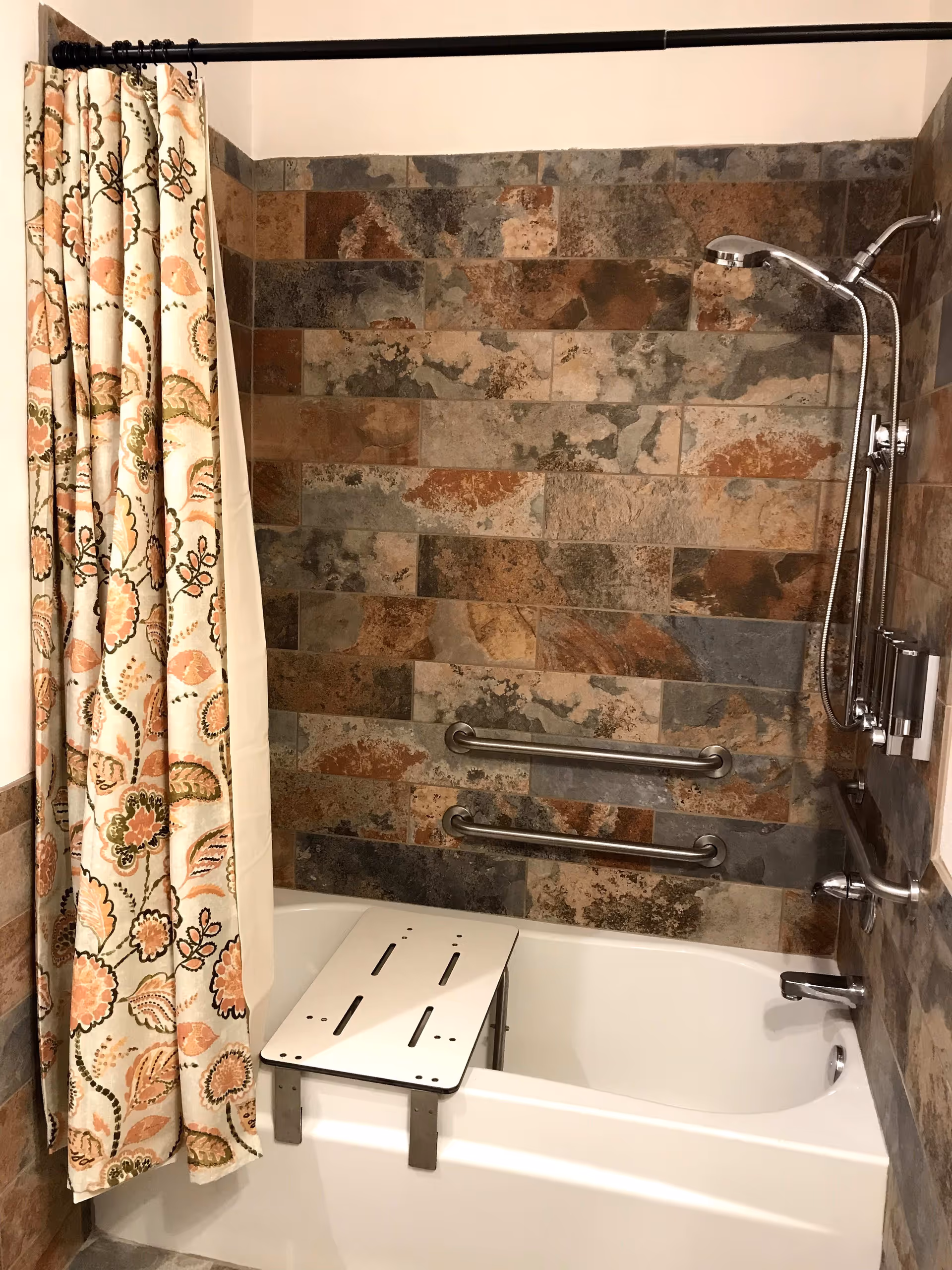 A bathtub with a mounted showerhead and two horizontal grab bars on the tiled wall. The tiles have a natural stone pattern in shades of brown, gray, and rust. A floral patterned shower curtain hangs on a rod to the left. A white bath bench is placed across the tub for seating.