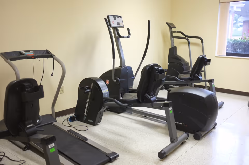 A small exercise room with various fitness equipment including a treadmill, a rowing machine, and two stationary recumbent bikes. The room has light yellow walls, a window with vertical blinds, and a light-colored floor.