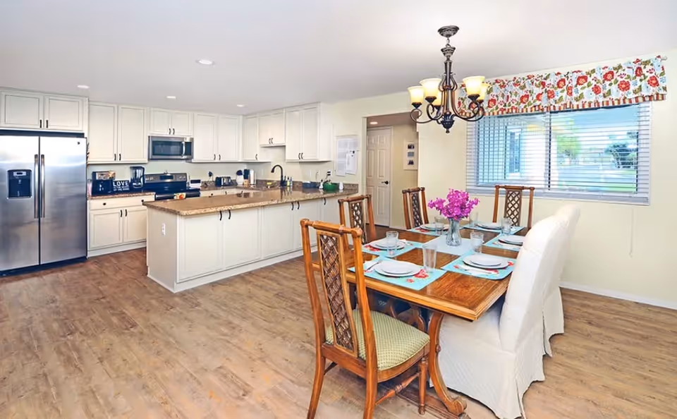 Bright and spacious kitchen and dining area with white cabinetry, stainless steel refrigerator, microwave, and stove. A wooden dining table is set with plates, glasses, and a vase of purple flowers under a decorative chandelier. A large window with floral valance lets in natural light.