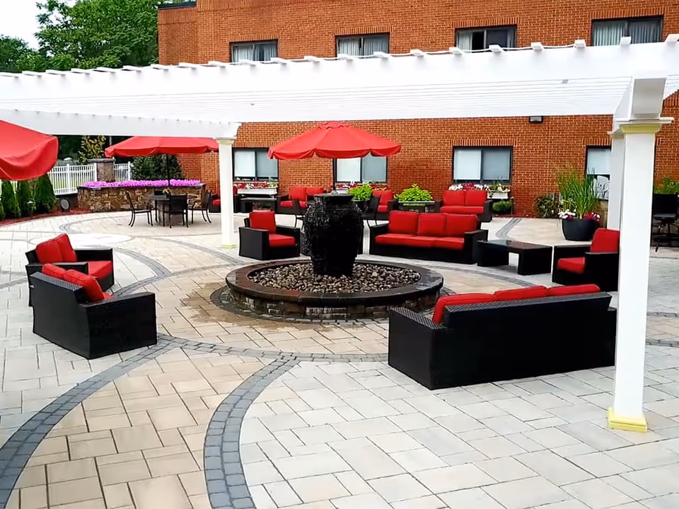 Outdoor patio area with black wicker furniture featuring red cushions arranged around a central water fountain. There are red umbrellas providing shade over some seating areas, a white pergola overhead, and a brick building in the background with windows and potted plants.