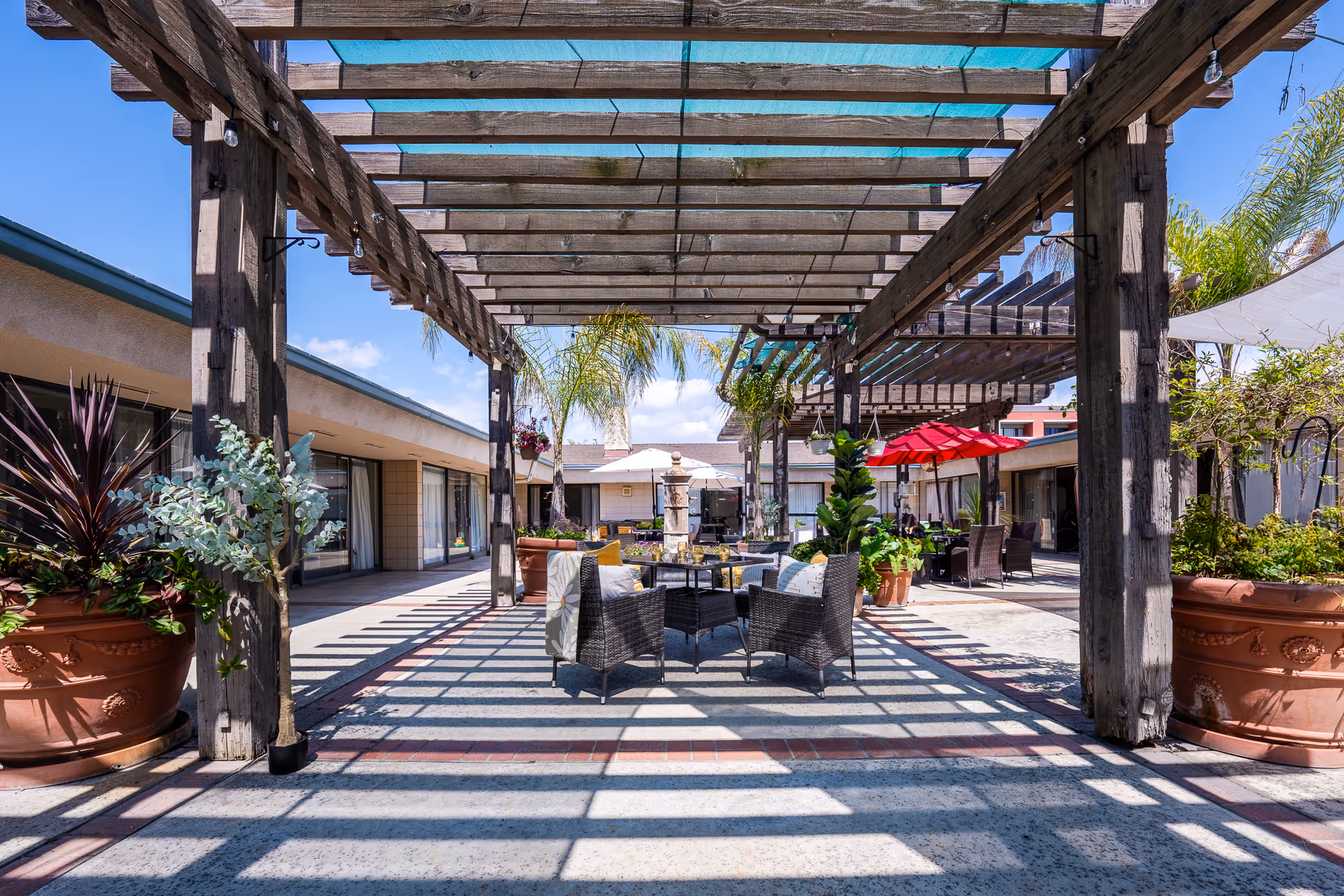 Outdoor patio area with wooden pergolas casting shadows on the ground, surrounded by large potted plants and palm trees. There are several seating arrangements with wicker chairs and tables, some shaded by umbrellas. The area is part of a building complex under a clear blue sky.
