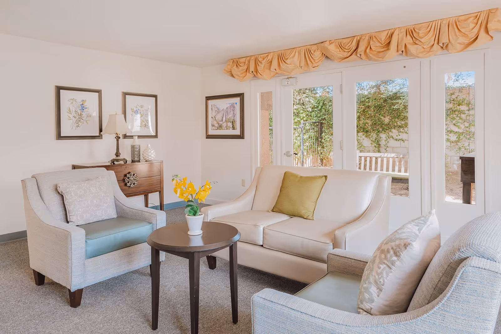 A bright and cozy living room with a white leather sofa adorned with a green pillow, two light blue armchairs with patterned cushions, and a round wooden coffee table with a small potted plant with yellow flowers. Behind the sofa, there are large glass doors leading to an outdoor area with greenery and a bench. The walls are decorated with framed artwork, and a wooden side table with a lamp and decorative items is placed against the wall.