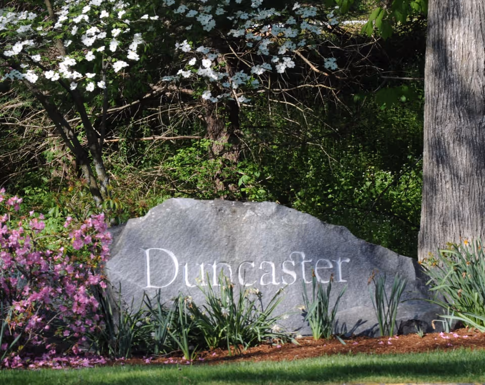 A large gray stone with the word 'Duncaster' engraved on it, surrounded by green plants, pink flowers, and a tree with white blossoms in a garden setting.