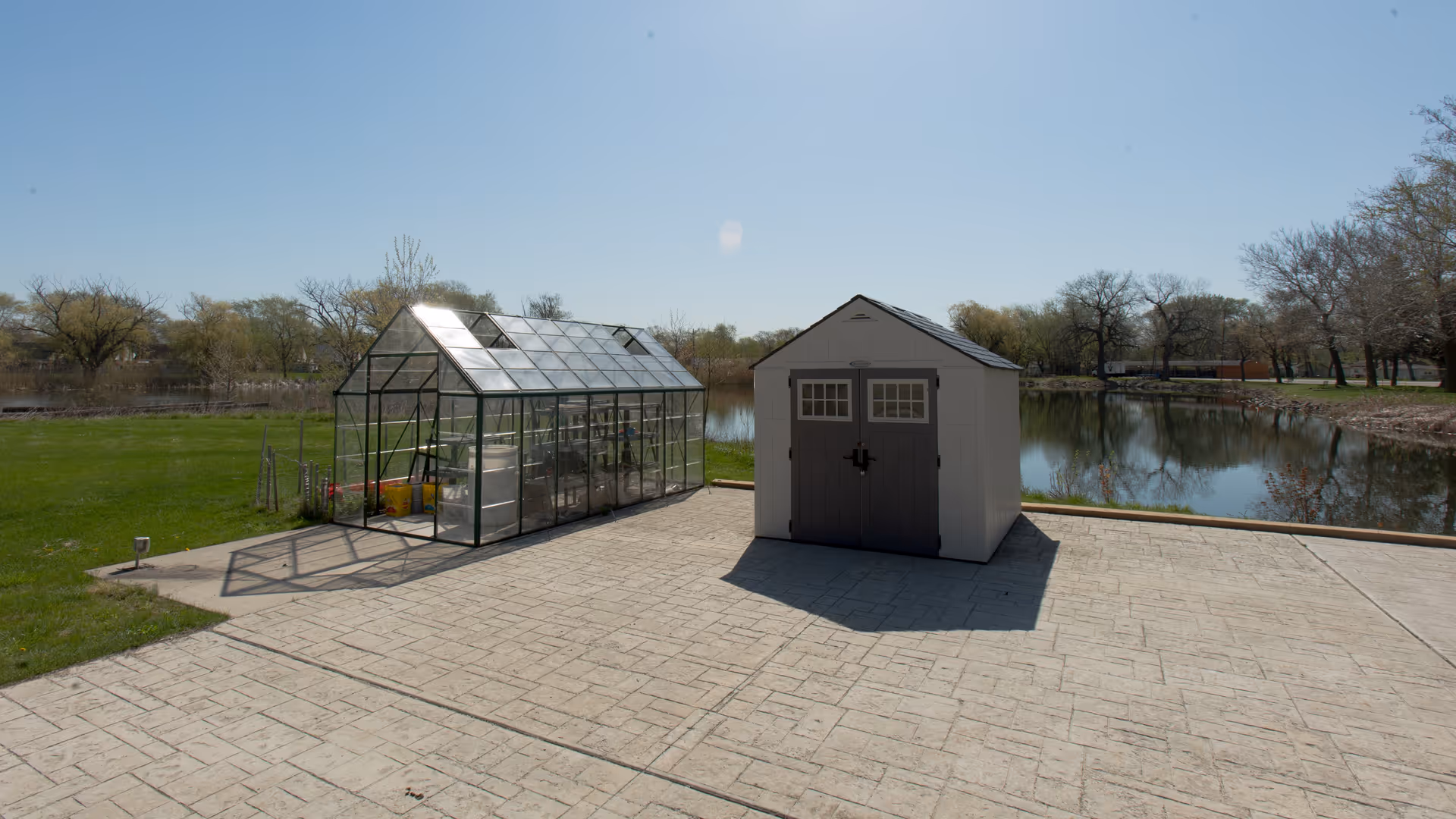 Outdoor scene showing a small glass greenhouse and a white storage shed with gray doors on a paved area next to a pond, surrounded by grass and trees under a clear blue sky.
