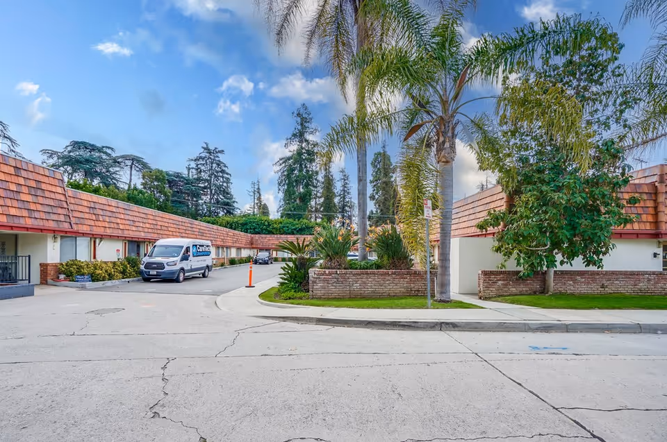 Front exterior of a low-rise senior care facility with a curved driveway, palm trees, a parked service van, and red-tiled roofs.