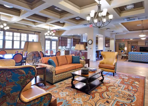 A spacious and well-lit living room area with patterned armchairs, an orange sofa with blue cushions, a wooden coffee table, and a large decorative rug. The room features coffered ceilings with chandeliers, large windows letting in natural light, and a kitchen area visible in the background.